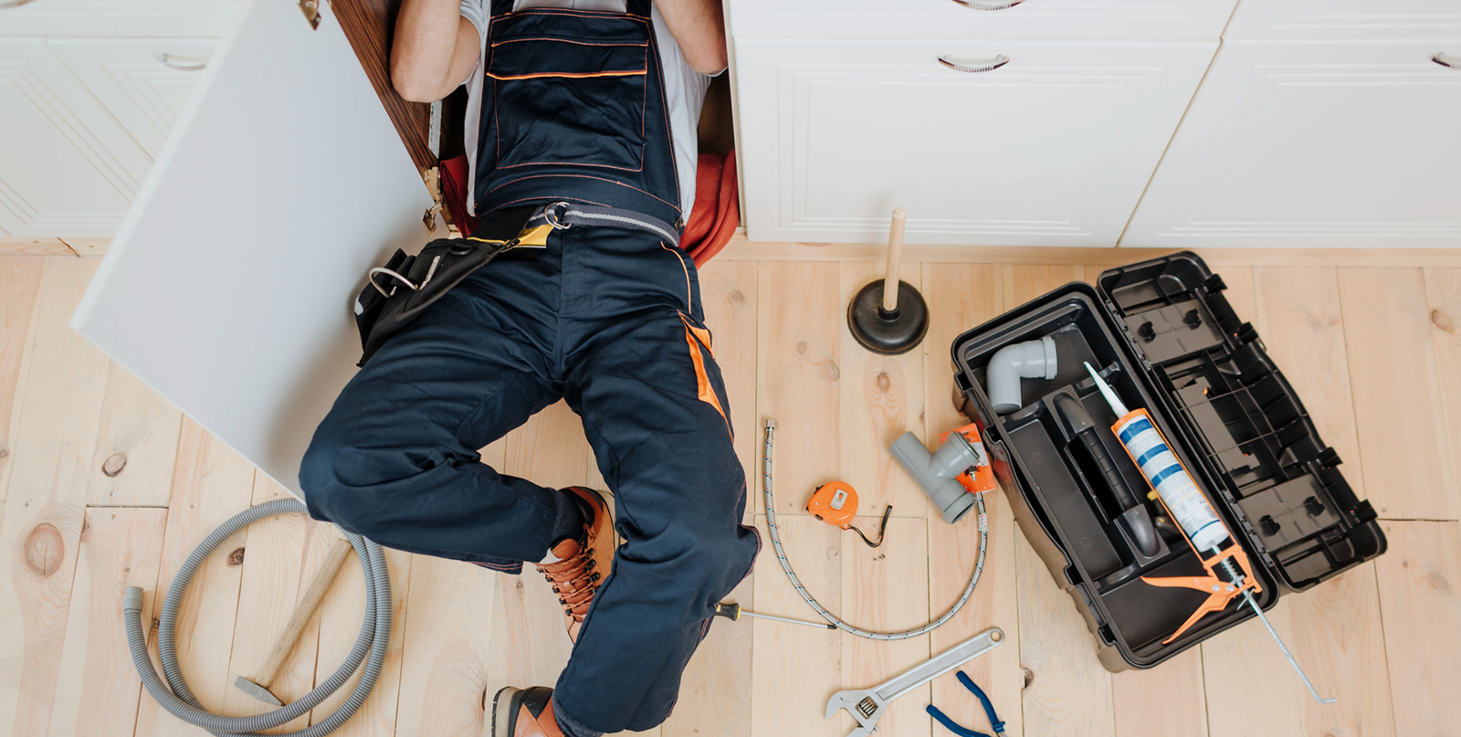 Man in uniform work in kitchen under sink. He repair water leaking. Hose and tools on floor. Opened toolbox